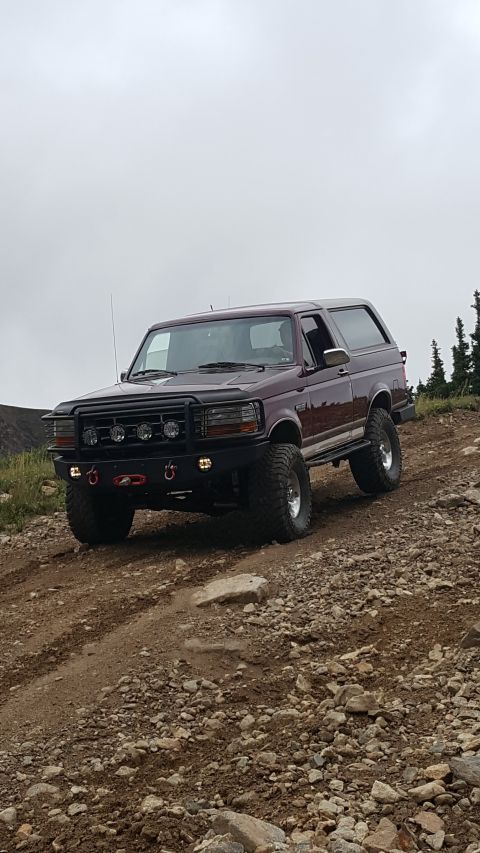 1996 Ford Bronco - A maroon 1996 Ford Bronco with off-road modifications is parked on a rocky, dirt trail with trees in the background.