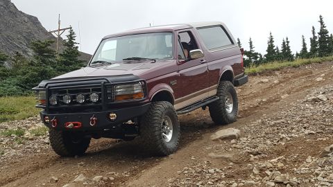 1996 Ford Bronco - A maroon 1996 Ford Bronco with a lifted suspension and off-road tires is driving up a rocky, mountainous dirt trail. The vehicle has a blac…