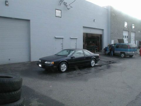 1990 Ford Thunderbird - A black 1990 Ford Thunderbird coupe is parked outside a commercial building with a blue van nearby.