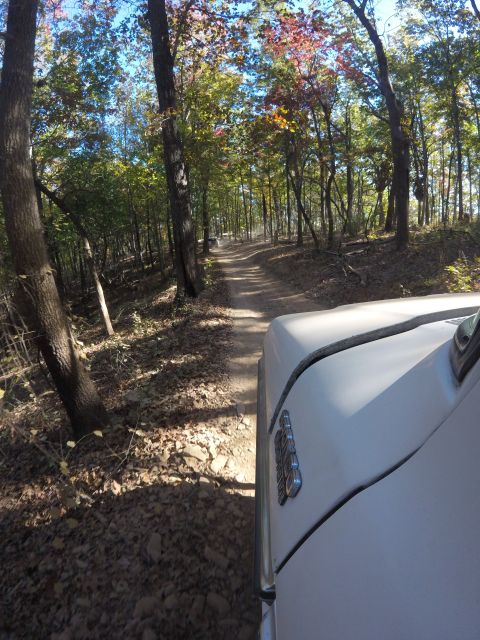 1979 Ford Bronco - The front right fender and hood of a white vehicle are visible in the foreground, driving on a dirt road through a wooded area with fall fo…