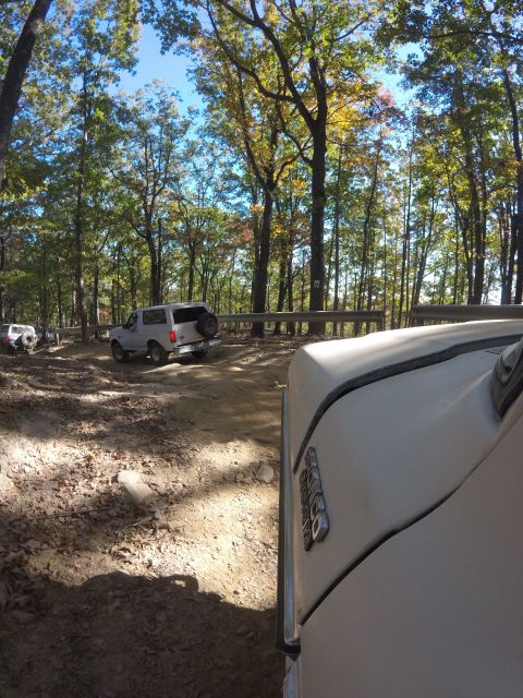 1979 Ford Bronco - A white Ford Bronco is parked on a dirt road surrounded by trees with autumn foliage. The perspective is from inside another vehicle, showi…