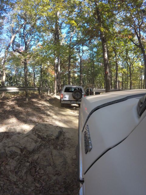 1979 Ford Bronco - The front of a white vehicle is shown from a low angle, driving on a dirt path through a wooded area. Another white SUV is visible ahead on…