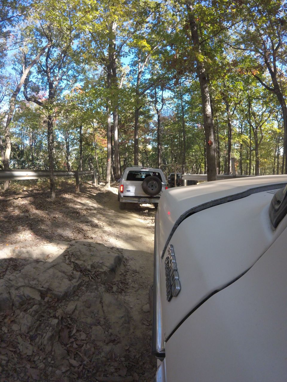 White vehicle driving on a dirt trail in a forest, with another SUV ahead.