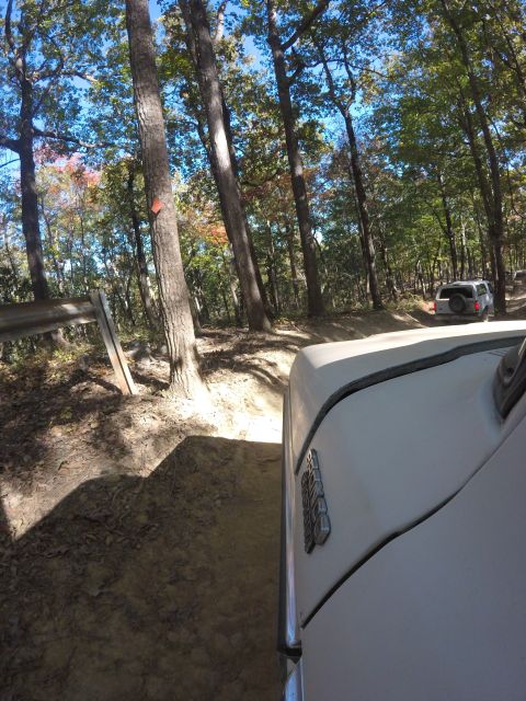 1979 Ford Bronco - The front passenger side of a white 1979 Ford Bronco is visible, driving on a dirt road through a forest with autumn foliage. Another white…