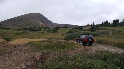 1996 Ford Bronco - A red 1996 Ford Bronco is parked on a dirt road in a mountainous, grassy landscape with scattered trees under a cloudy sky. The vehicle is…