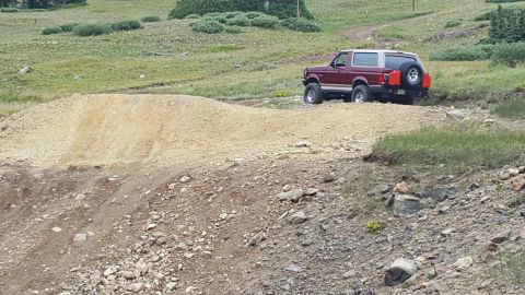 1996 Ford Bronco - A maroon and tan 1996 Ford Bronco is parked on a dirt road in a grassy, mountainous area. The vehicle has two red jerry cans mounted on the…