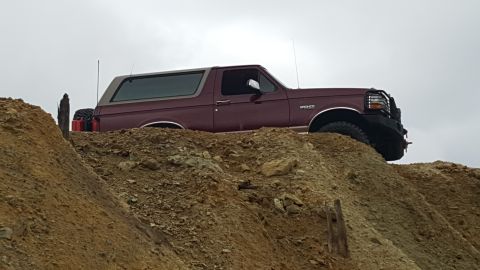 1996 Ford Bronco - A maroon Ford Bronco is parked on a dirt mound with a spare tire and gas can visible behind it. The vehicle is viewed from the side, with a…