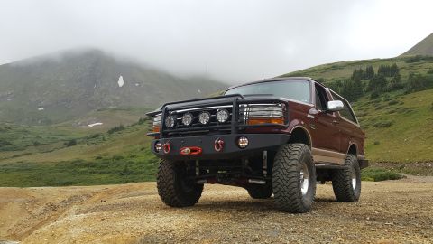 1996 Ford Bronco - A lifted, maroon 1996 Ford Bronco with an aftermarket front bumper and grille guard is parked on a dirt road in a mountainous, foggy landsc…