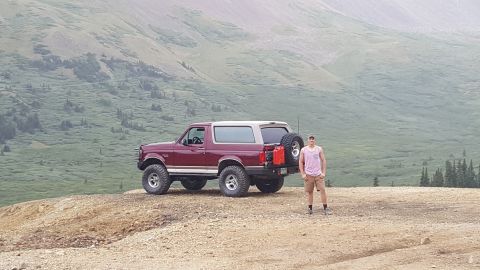 1996 Ford Bronco - A man stands next to a maroon Ford Bronco with a tan top parked on a dirt overlook with a mountainous, tree-covered landscape in the backgr…