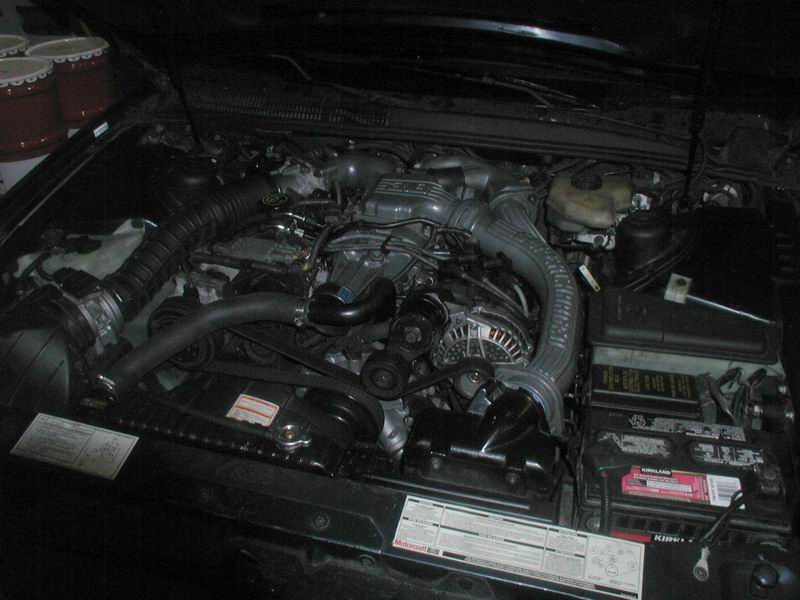 Engine bay of a dark car showing V6 engine, serpentine belt, alternator, and Kirkland battery