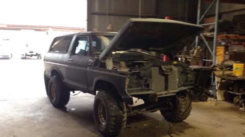 1979 Ford Bronco - A dark-colored, lifted Ford Bronco is parked inside a garage with its hood open, revealing the engine bay. The vehicle has large off-road t…
