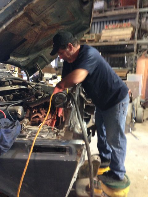 1979 Ford Bronco - A man wearing a black baseball cap and blue jeans works on the engine of a vehicle with its hood open. He is standing on two yellow buckets…