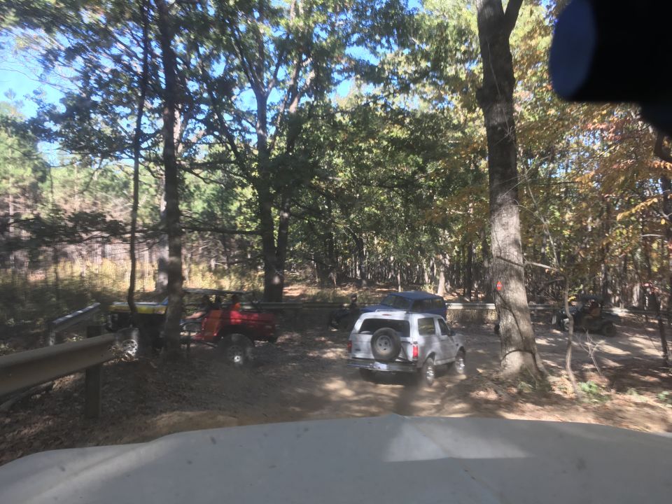 White Ford Bronco driving on a dirt path in a wooded area with other off-road vehicles.