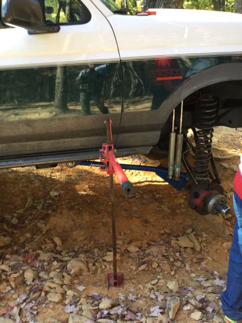 1979 Ford Bronco - A red Hi-Lift jack is positioned under the frame of a white and dark green Ford Bronco, lifting the front passenger side of the vehicle. Th…