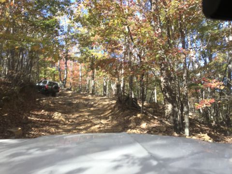 1979 Ford Bronco - A dark-colored SUV is driving up a steep, rocky, off-road trail through a forest with autumn foliage. The vehicle is viewed from behind and…