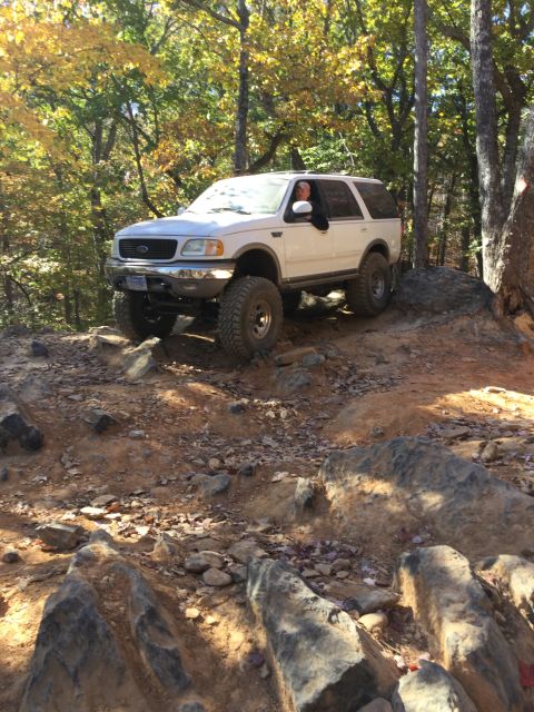 1979 Ford Bronco - A white Ford Expedition is driving off-road on a rocky, dirt trail surrounded by trees with fall foliage. The driver is visible through the…