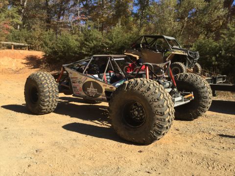 1979 Ford Bronco - A custom-built, off-road buggy with large tires is parked on a dirt path, with a utility vehicle on a trailer behind it. The buggy has a ro…