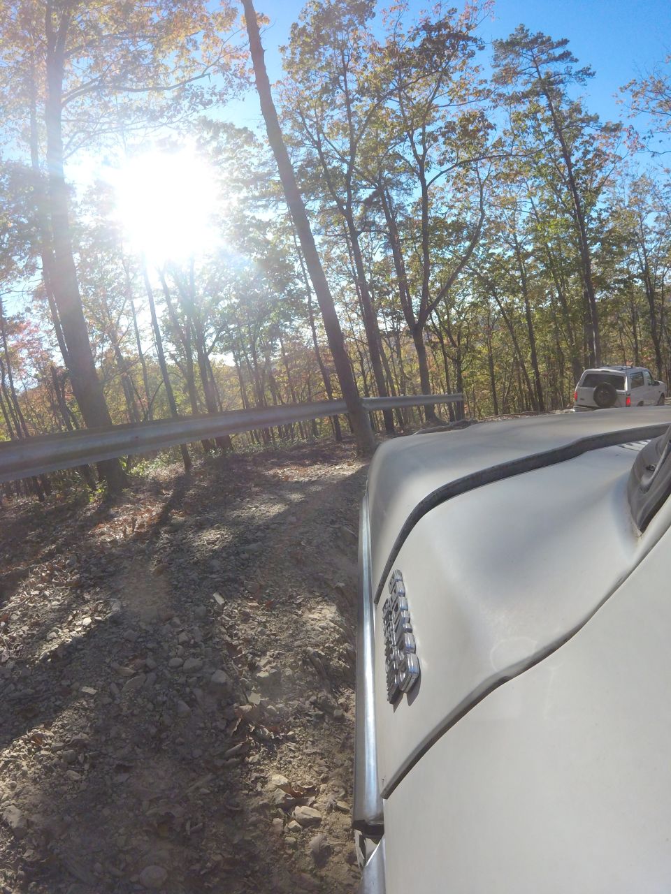 Light-colored vehicle driving on a gravel road in a forest with sun shining through trees.