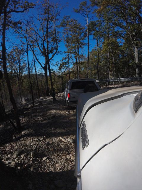 1979 Ford Bronco - The front fender and hood of a white vehicle are in the foreground, with a silver pickup truck driving on a rocky, wooded trail behind it.…
