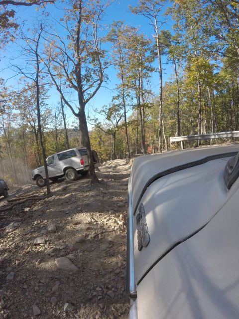 1979 Ford Bronco - A white Ford Bronco is navigating a rocky, wooded trail. The photo is taken from the perspective of another vehicle, showing the side of it…