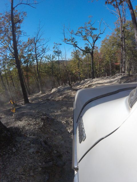 1979 Ford Bronco - The front corner of a white vehicle is visible, driving on a rocky, off-road trail through a forest with trees showing fall colors.