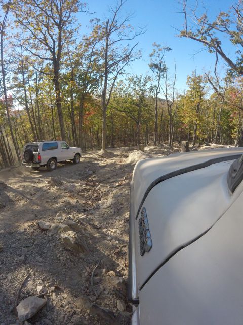 1979 Ford Bronco - A white 1979 Ford Bronco is parked on a rocky, off-road trail surrounded by trees with autumn foliage. The camera is positioned low, showin…