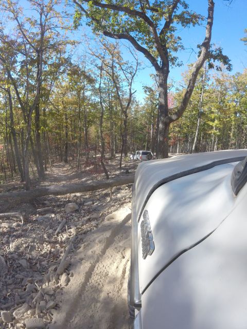 1979 Ford Bronco - A white vehicle is driving on a dirt and rocky path through a forest with trees showing fall colors. The front right fender and hood of the…