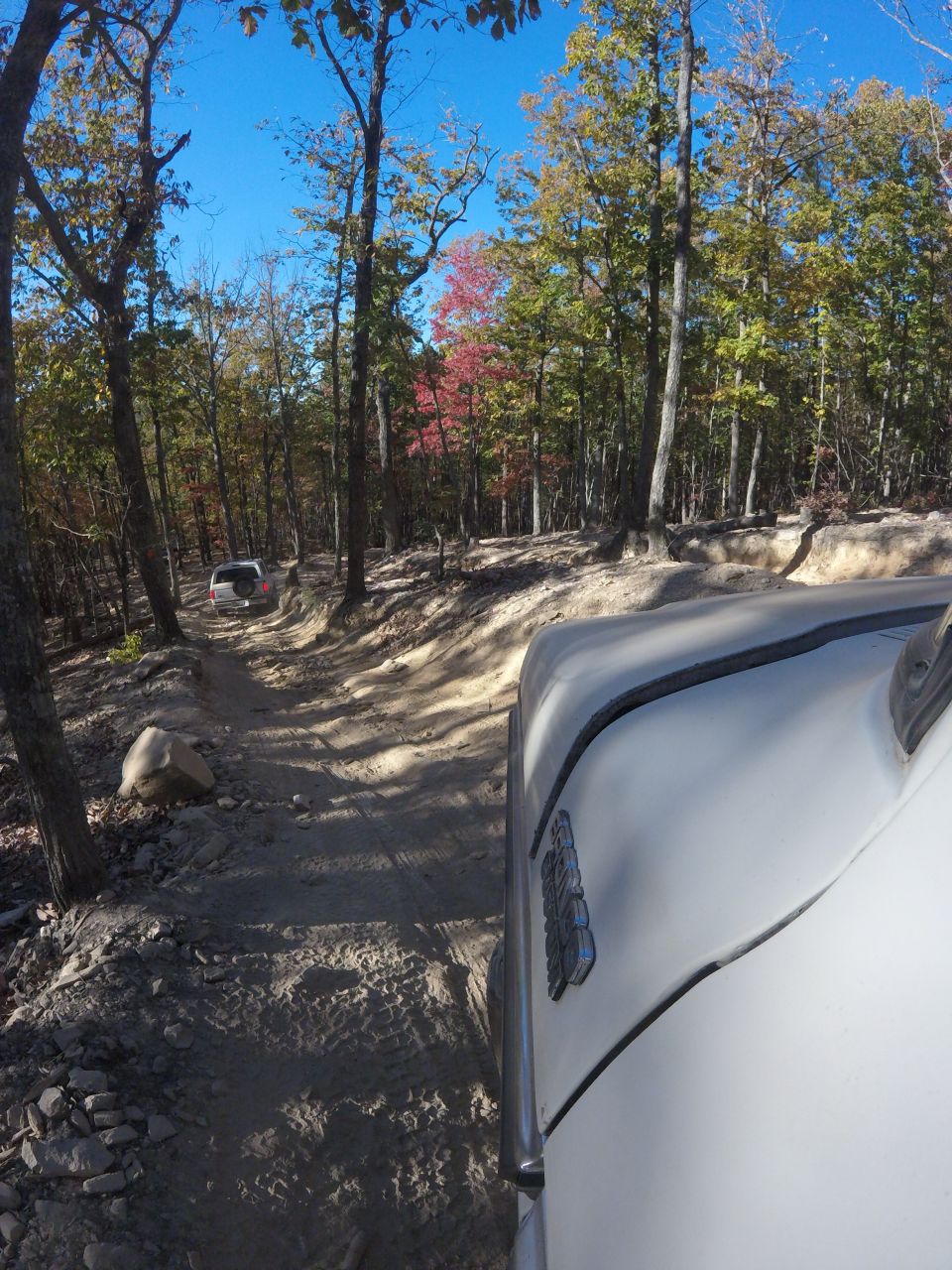White vehicle on a dirt road in a forest with autumn leaves.