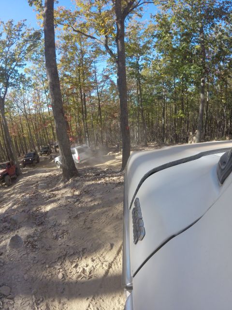 1979 Ford Bronco - A white Ford Bronco is seen from the side, driving on a dirt road through a forest with other off-road vehicles visible in the background.