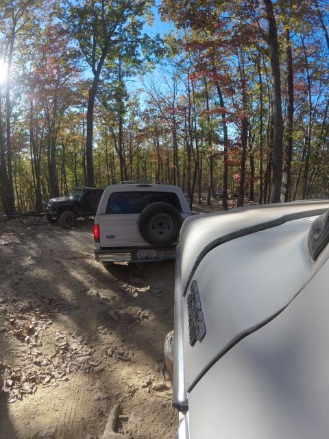 1979 Ford Bronco - A white Ford Bronco is parked on a dirt trail surrounded by trees with autumn foliage. Another black Jeep is visible behind it.