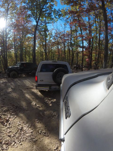1979 Ford Bronco - A white Ford Bronco is parked on a dirt trail in a forest with autumn foliage. Another off-road vehicle is visible in the background.