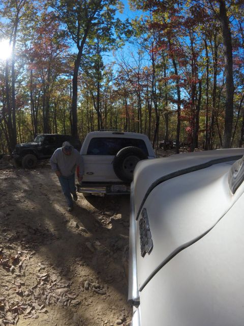 1979 Ford Bronco - A man walks past a white Ford Bronco with a spare tire mounted on the back, parked on a dirt trail surrounded by trees with autumn foliage.…