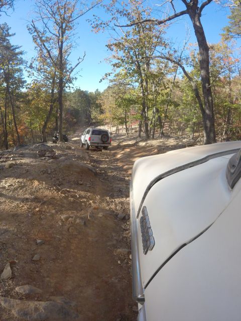 1979 Ford Bronco - A white Ford Bronco is driving on a rocky, dirt trail through a wooded area with fall foliage. The photo is taken from the perspective of a…