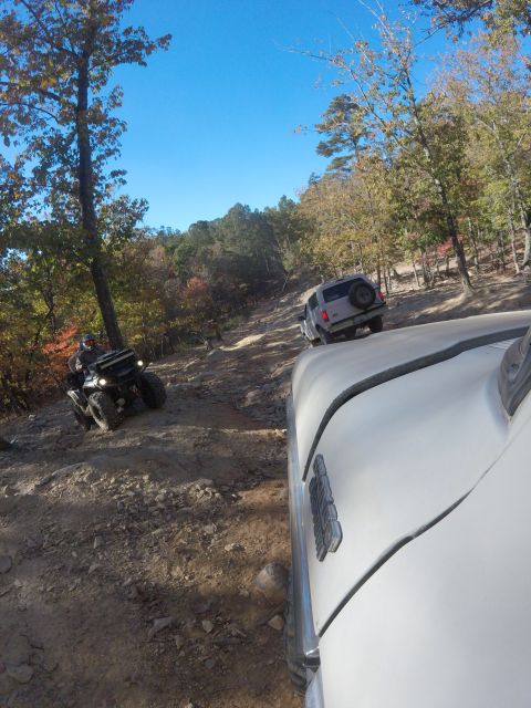 1979 Ford Bronco - A white SUV is driving on a rocky, wooded trail, with a black ATV visible ahead and to the left. The perspective is from another vehicle, s…
