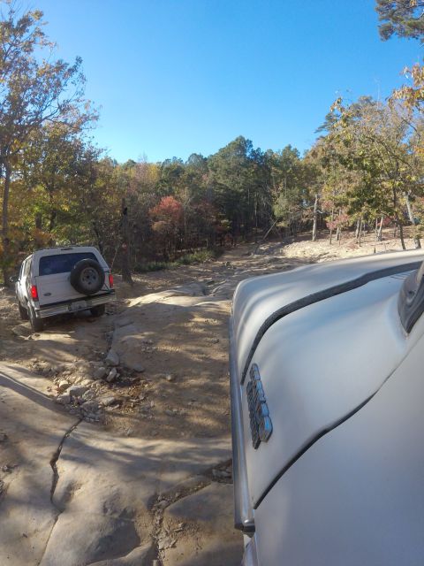 1979 Ford Bronco - A white SUV with a spare tire mounted on the back is driving on a rocky, off-road trail. The perspective is from another vehicle following…