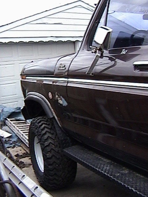 1995 Ford Bronco - Tight low-angle view of the driver-side front fender and door of a dark brown late-1970s Ford truck parked beside a white garage, showing a…