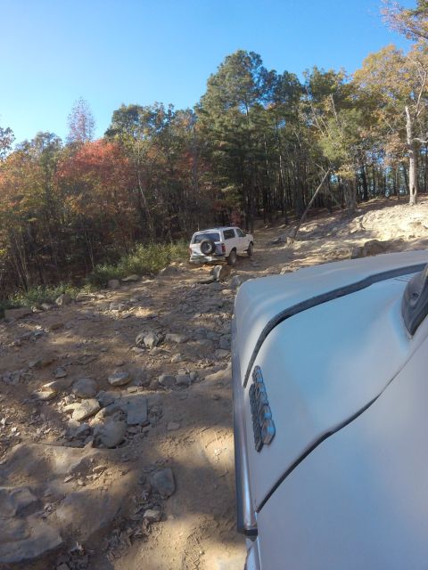 1979 Ford Bronco - A white SUV is driving on a rocky, dirt road through a forest with autumn foliage. The front right fender and door of another white vehicle…