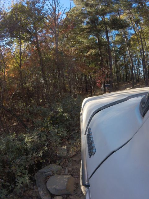 1979 Ford Bronco - The right side of a white vehicle is visible, driving on a rocky, wooded trail during autumn. The vehicle's fender has a chrome emblem.