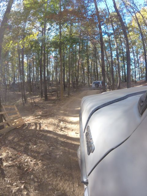 1979 Ford Bronco - The front corner of a light-colored vehicle is visible in the foreground, driving on a dirt road through a forest. Another white SUV is vis…