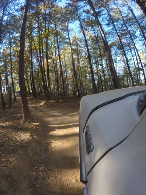 1979 Ford Bronco - A white vehicle is driving on a dirt road through a forest during the daytime. The front passenger side of the vehicle is visible, showing…