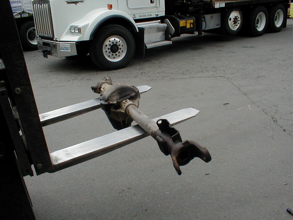 Front axle and differential balanced on forklift forks in front of a white Kenworth flatbed truck