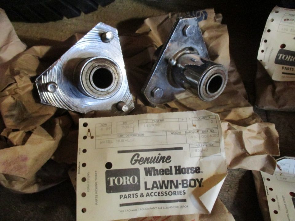 Two metal wheel hubs with bearings, one triangular and one with a bracket, next to Toro and Lawn-Boy parts packaging