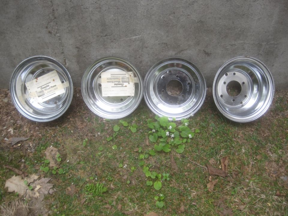 Four chrome wheels with labels on two of them, resting on grass in front of a concrete wall