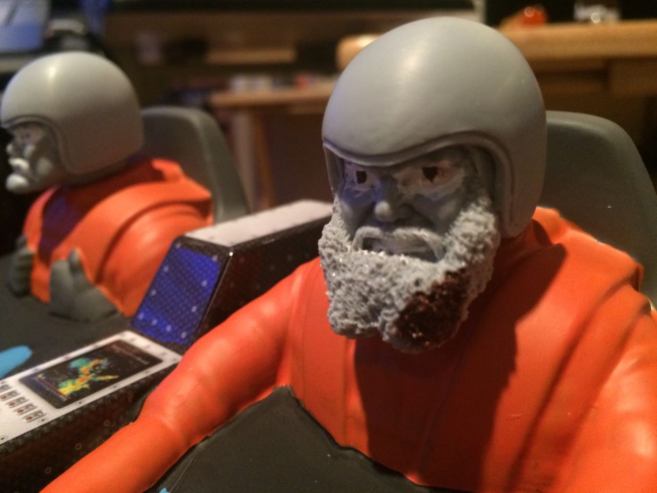 Two toy astronaut figures with helmets and orange suits sit in a cockpit