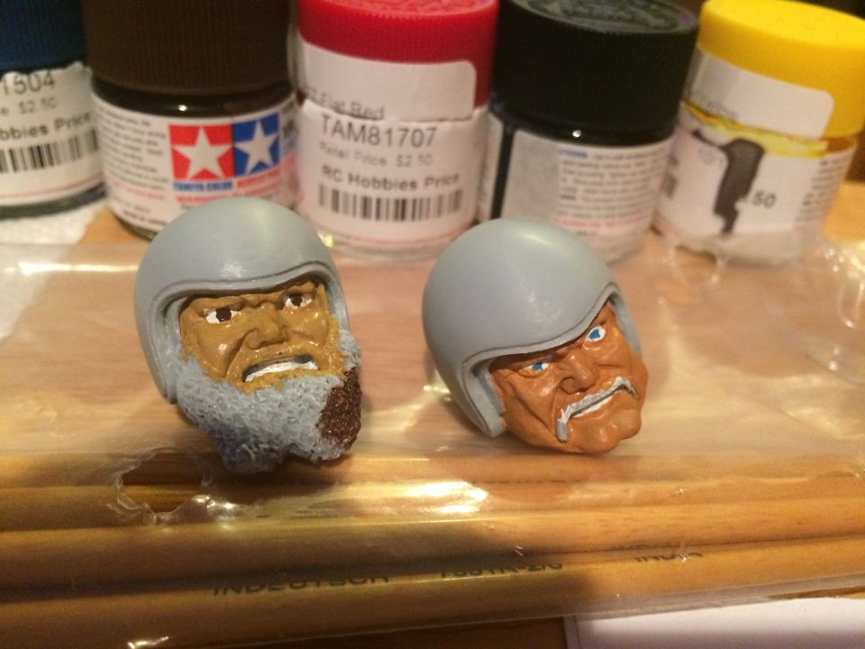Two unpainted model kit heads with helmets sit in front of several small paint bottles