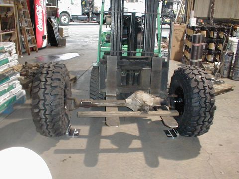 1983 Ford Bronco - A complete solid front axle assembly with mud-terrain tires mounted on both ends, lifted on the forks of a green forklift inside a warehous…