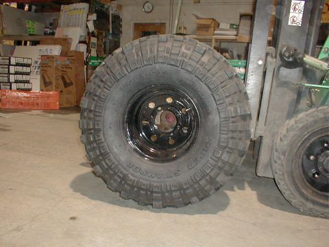 1983 Ford Bronco - A Super Swamper TSL/SX mud tire mounted on a black 8-lug steel wheel, standing on a warehouse floor next to a green forklift, with the side…