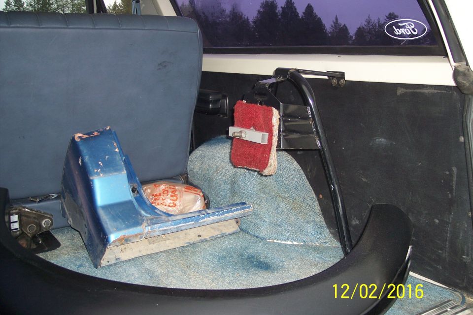 Interior rear of a vehicle with blue seats, a blue plastic part on the floor, and a red sponge hanging from a bar. Ford logo on rear window