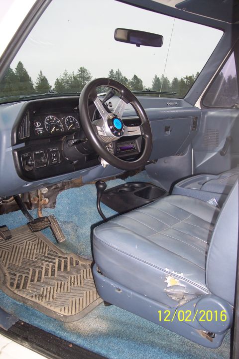 1990 Ford Bronco - The interior of a vintage Ford Bronco shows a blue dashboard, aftermarket steering wheel, and worn blue vinyl seats. A rubber floor mat cov…