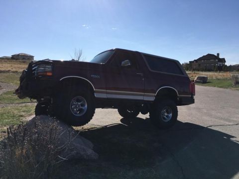 1996 Ford Bronco - A maroon 1996 Ford Bronco is positioned on a rock with its front tires elevated. The SUV is parked on a paved surface next to a grassy area…